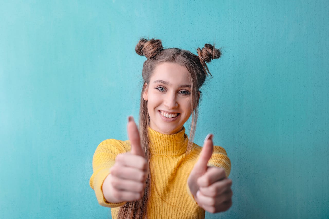 Happy woman in yellow giving thumbs up against a teal background, expressing positivity.